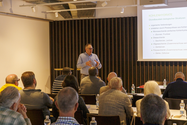 Ein Mann im Hemd präsentiert einen Macardo Brenn-Kurs vor Publikum. Eine Leinwand zeigt Informationen über Kohlenhydrate. Mehrere Personen sitzen an Tischen mit Wasserflaschen und Notizen. Sie hören dem Vortrag aufmerksam zu.
